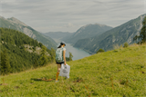 Frau mit Rucksack wandert mit einem Clean Up-Sack über eine Bergwiese mit Blick auf den Achensee
