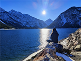 Frau in Winterkleidung sitz auf einem Stein, vor ihr ein grosser See mit verschneiten Bergen dahinter, Sonne steht am Himmel